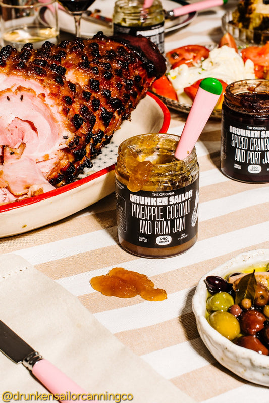 Drunkern Sailor pineapple coconut and rum jam jar on a table with a sliced ham and other food items.