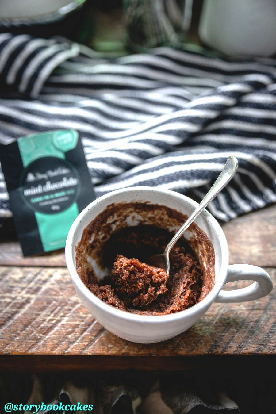 Mug cake in a white cup with a spoon, next to a packet of mix on a striped cloth.