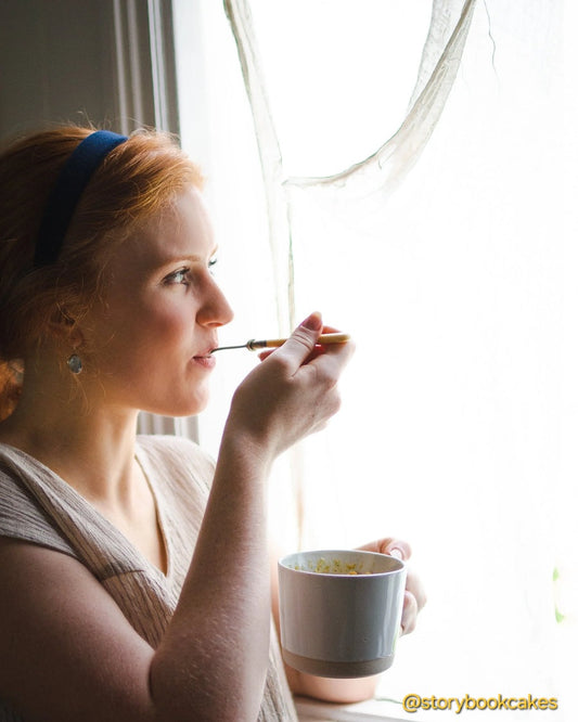 Woman applying makeup with a brush and looking out a window, holding a mug.