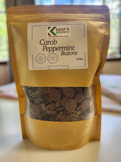 A stand-up pouch of Krisp n Krunch Carob Peppermint Buttons, showing the dark brown chocolate-like buttons through a clear window in the lower half, set on a light surface with a blurred indoor background.