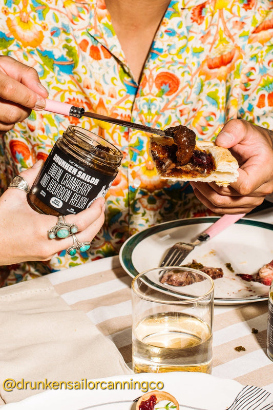 Person spreading Drunken Sailor Relish on a slice of bread with a jar of jam and a glass of water on a table.
