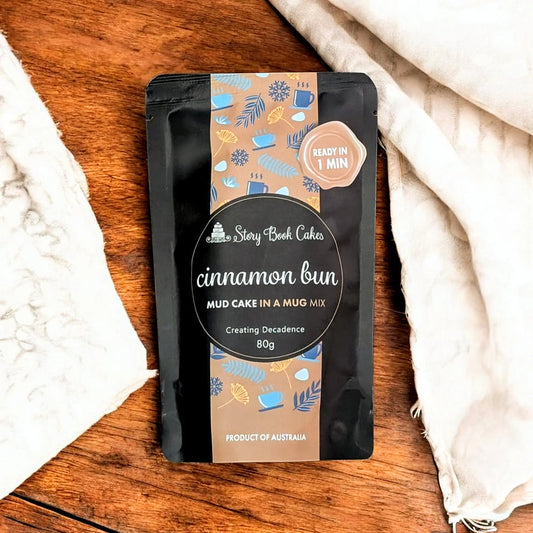 Packaged cinnamon bun mud cake in a mug mix on a wooden surface with a white cloth.
